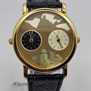 May include: Gold-coloured Geneva quartz watch with a black leather strap. The watch face has two dials, one with Arabic numerals and the other with Roman numerals. The watch has a world map design.