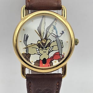 May include: A gold-coloured wristwatch with a brown leather strap. The watch face displays a cartoon image of Wile E. Coyote. The watch has a classic design with a round face and a brown leather strap.