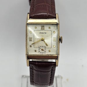 May include: A vintage gold-toned LeCoultre wristwatch with a rectangular face and a brown leather strap. The watch face features gold hands and hour markers, with the brand name and Roman numerals. The strap is a dark brown colour.