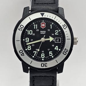May include: A black and silver Swiss Army watch with a black leather band. The watch face has white numbers and a red cross logo. The band is a braided style.