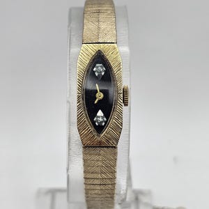 May include: A vintage gold-coloured wristwatch with a black oval face. The watch features two small, sparkling accents and a gold-coloured band with a textured design. The watch is displayed on a clear stand against a white background.
