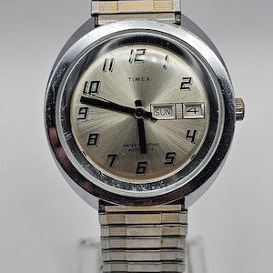 May include: Silver-toned Timex watch with a silver dial, featuring black hands and numerals. The watch displays the day and date. The watchband is a silver and gold-toned expansion band. The watch reads "TIMEX" and "WATER RESISTANT AUTOMATIC".