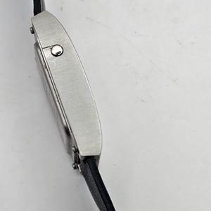 以下が含まれることがあります： 黒い革バンドが付いた、銀色の長方形の腕時計。時計の文字盤には小さな円形のディテールがあります。バンドは小さな金属ピンで時計に取り付けられています。