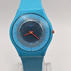 Puede incluir: Un reloj Swatch Swiss con correa y caja azul claro. La esfera del reloj es azul oscuro con un anillo azul más claro y agujas rojas. El reloj se muestra sobre un soporte transparente.