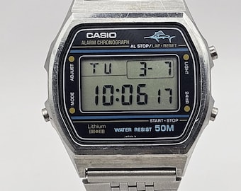 Puede incluir: Reloj digital Casio plateado con una esfera rectangular que muestra la hora 10:06:17. El reloj tiene una correa de metal y varios botones de función. La esfera del reloj incluye el texto "CASIO ALARM CHRONOGRAPH".