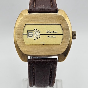 May include: Vintage Lucerne Digital watch with a gold-tone case and a brown leather strap. The watch face displays the time digitally with the brand name and "SWISS" at the bottom. The watch is displayed on a clear stand.
