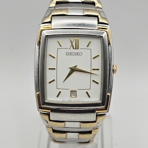 May include: A Seiko wristwatch with a rectangular white face, gold-tone accents, and a silver-tone band. The watch face displays the number 16 and the Seiko logo. The watch is displayed on a clear stand.