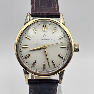 Puede incluir: Un reloj de pulsera dorado con una esfera blanca y una correa de cuero negra. La esfera del reloj tiene marcadores de hora en números romanos negros y el texto "ETERNA-MATIC".