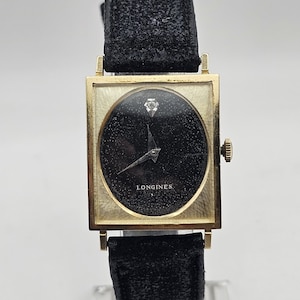 May include: A vintage Longines watch with a square gold-tone case and a black dial. The watch features a small diamond at the 12 o'clock position and black leather straps. The watch is displayed on a clear stand.