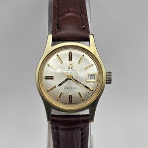 Pode incluir: Um relógio de pulso automático Omega Geneve dourado com mostrador branco e pulseira de couro marrom.