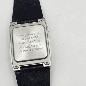 以下が含まれることがあります： 黒い革バンドが付いた長方形のシルバーの腕時計。文字盤には「Timepiece by Charles S. Coster, Designed in USA, Assembled in China, Time by Design, U.S. Patent D 348403」と書かれています。