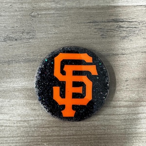 May include: A round, black glittered emblem featuring the orange "SF" logo of the San Francisco Giants baseball team. The logo is bold and centered on the emblem.