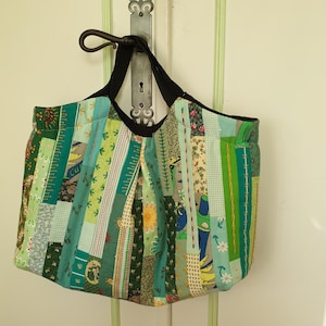 Può includere: Una borsa tote patchwork con manico e finiture nere. La borsa è realizzata con vari tessuti verdi e blu, cuciti insieme a strisce verticali. La borsa è appesa a una porta bianca.