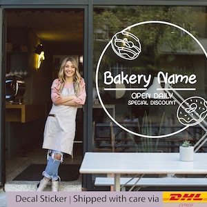 May include: A white circle decal sticker with the text "Bakery Name", "Open Daily", and "Special Discount". The sticker also features two outline drawings of pastries. The sticker is on a glass door of a bakery.