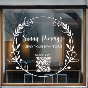 Könnte beinhalten: Ein weißer Vinyl-Fensteraufkleber mit einem kreisförmigen Blumenkranz-Design. Der Text lautet Sunny Pimmy.co, SEND YOUR INFO. TO US und SCAN HERE, ergänzt durch einen quadratischen QR-Code. Das Design ist auf einer Glasfläche mit Holzrahmen angebracht.