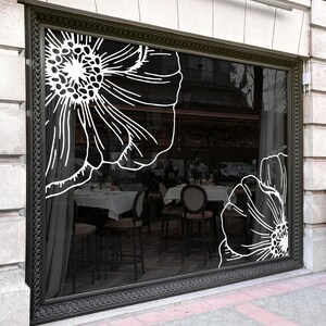 Op de afbeelding: Een groot rechthoekig etalageraam met een donker metalen frame, voorzien van twee witte lijntekening-bloemstickers op het glas. Het interieur toont een restaurant met wit gedekte tafels en donkerhouten stoelen.