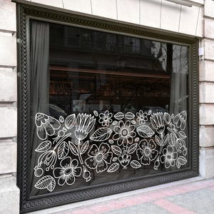 Peut inclure: Une grande vitrine rectangulaire encadrée de métal sombre, ornée d'un autocollant en lignes blanches représentant diverses fleurs et feuilles sur la vitre. Le motif floral couvre la moitié inférieure de la fenêtre, contrastant avec l'intérieur.