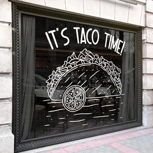 May include: A window display with the text "IT'S TACO TIME!" above a white line drawing of a taco with a lime slice. The window is framed in black and has a dark interior with tables and chairs visible.