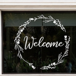 May include: A white vinyl window decal featuring the word Welcome in a cursive script font, centred within a circular wreath design composed of thin branches, leaves, and small tulip-like flower buds, applied to a glass window pane.