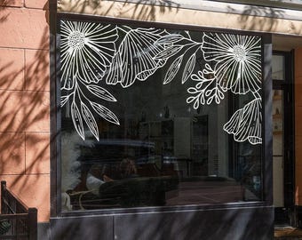 以下が含まれることがあります： 店舗のショーウィンドウに貼られた白い線画のフローラルデカール。ガラスの上部コーナーに配置された大きな様式化された花と葉の枝のデザインが、鮮明で大胆な白い輪郭を持つ装飾的なボーダーを作り出しています。