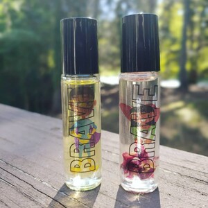 May include: Two clear glass roller bottles with black caps. Both bottles have a label that says "BRAVE" in black letters. One bottle has a colorful illustration of a superhero, and the other bottle has a pink flower inside.