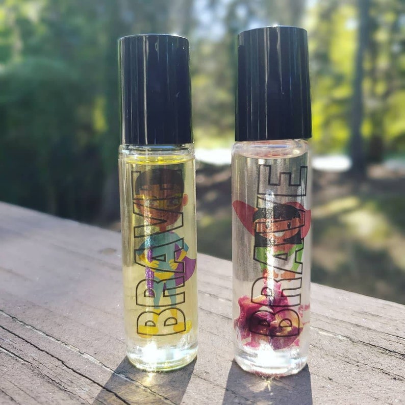 May include: Two clear glass roller bottles with black caps. Both bottles have a label that says "BRAVE" in black letters. One bottle has a colorful illustration of a superhero, and the other bottle has a pink flower inside.