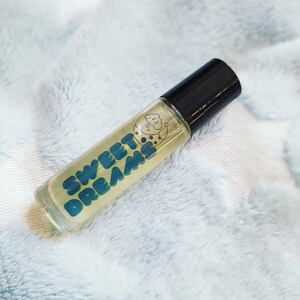 May include: A clear glass roller bottle with a black cap. The bottle is filled with a yellow liquid and has a blue and white label that says "SWEET DREAMS" with a crescent moon and a cloud graphic.