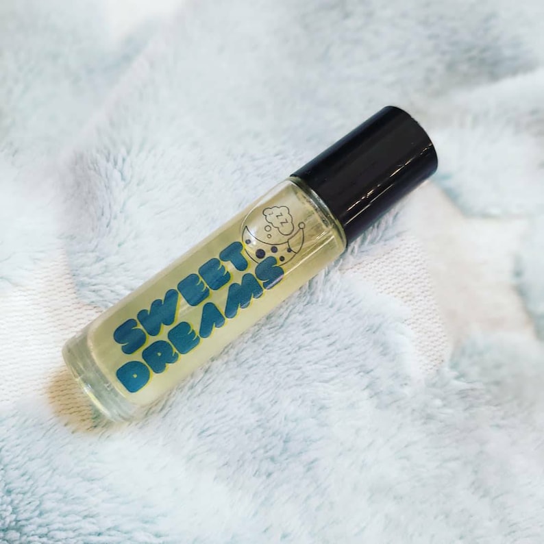 May include: A clear glass roller bottle with a black cap. The bottle is filled with a yellow liquid and has a blue and white label that says "SWEET DREAMS" with a crescent moon and a cloud graphic.