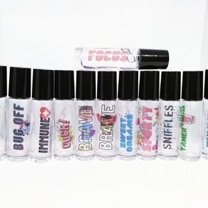 May include: A set of 12 clear glass roller bottles with black caps and colorful labels. The labels feature various phrases and images, including "Happy", "Bug Off", "Immune", "Ouch!", "Brave", "Sweet Dreams", "Sporty", "Sniffles", "Tamer", "Tummy", and "Trouble".