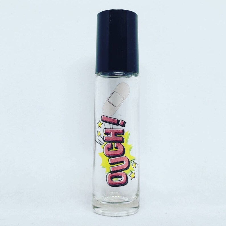 May include: A clear glass roller bottle with a black lid and a cartoon-style label that says "OUCH!" in pink letters with a yellow background. A white bandage is on the label.