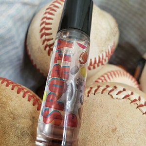 May include: A clear glass roll-on bottle with a black cap. The bottle has a label with the word "Sporty" in red and blue letters. The label also features a variety of sports-themed graphics, including baseballs, footballs, and basketballs.