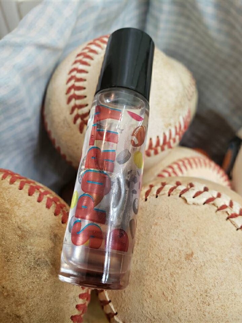 May include: A clear glass roll-on bottle with a black cap. The bottle has a label with the word "Sporty" in red and blue letters. The label also features a variety of sports-themed graphics, including baseballs, footballs, and basketballs.
