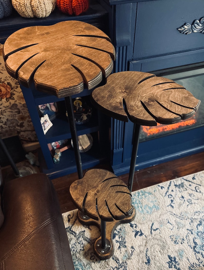 May include: Three tiered plant stand made of wood and metal. Each tier is shaped like a monstera leaf and is painted a dark brown color. The stand is supported by three black metal legs.
