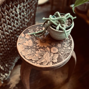 May include: A small wooden stool with a floral design carved into the top. A potted succulent plant sits on top of the stool.