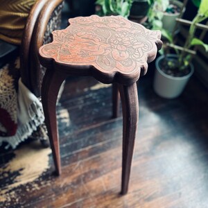 May include: A small, brown wooden side table with a decorative, floral-patterned top. The table has three curved legs and stands on a wooden floor. The table's top is approximately 12 inches in diameter.