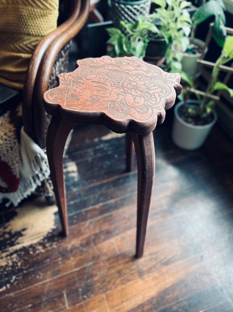 May include: A small, brown wooden side table with a decorative, floral-patterned top. The table has three curved legs and stands on a wooden floor. The table's top is approximately 12 inches in diameter.