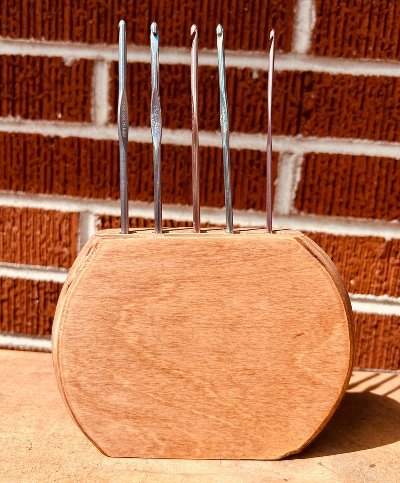 May include: A wooden stand with six metal crochet hooks. The stand is brown and has a rounded shape. The hooks are silver and have different sizes.