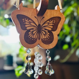 May include: Wooden butterfly wind chime with a detailed butterfly engraving. The wind chime features clear crystal accents that dangle below the wooden butterfly. The wind chime is suspended by a cream-colored rope.