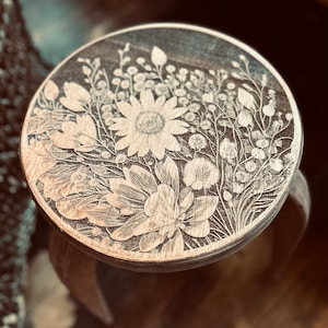 May include: A wooden stool with a round top featuring a laser-etched floral design. The design includes a large flower with multiple petals and smaller flowers and leaves.