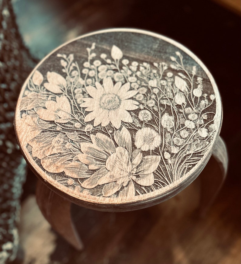 May include: A wooden stool with a round top featuring a laser-etched floral design. The design includes a large flower with multiple petals and smaller flowers and leaves.