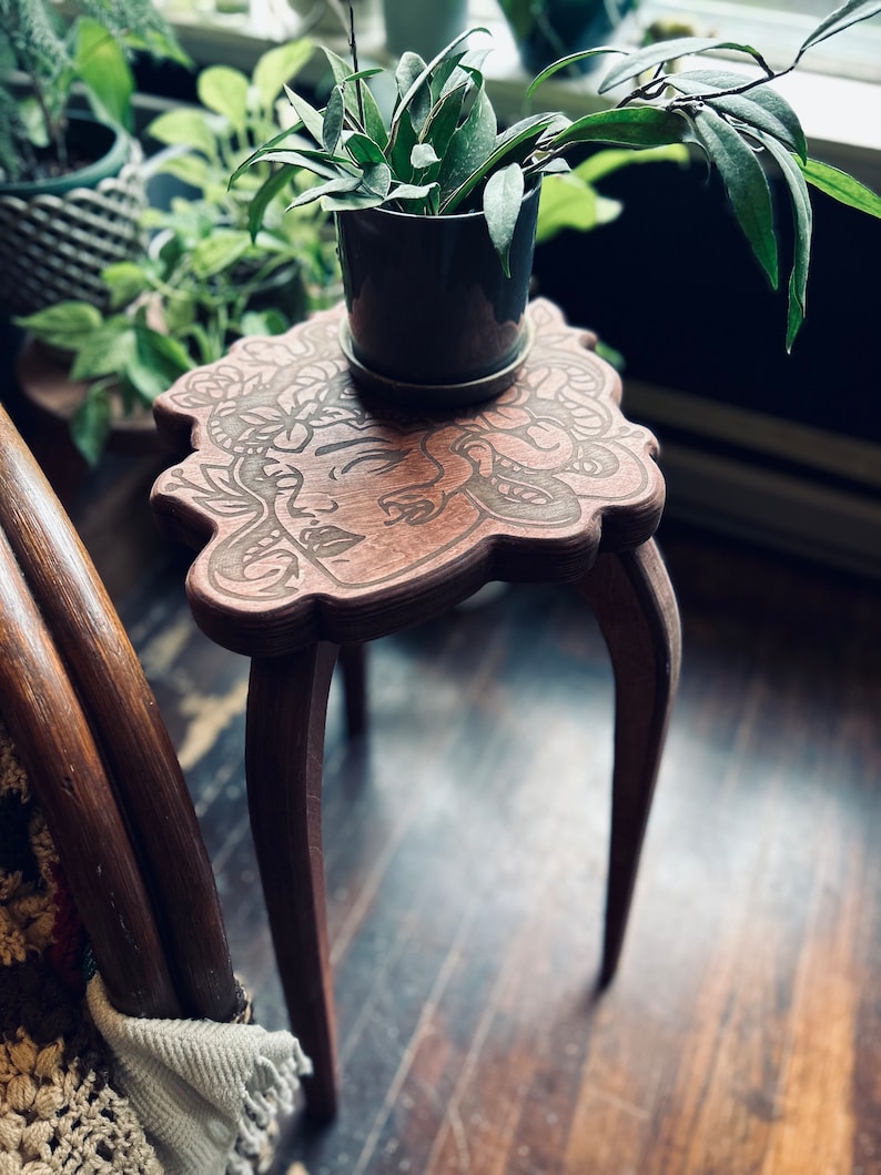 May include: A small, brown wooden plant stand with a carved floral design and three curved legs. A potted plant with green leaves sits on top. The stand is placed on a dark wooden floor.