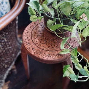 Könnte beinhalten: Ein Holzhocker mit einem geschnitzten Mandala-Design auf der Oberseite. Eine Topfpflanze mit grünen Blättern steht auf dem Hocker.
