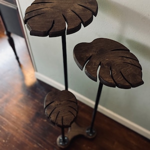 May include: A three-tier plant stand made of black metal pipes and dark brown wooden shelves shaped like monstera leaves.