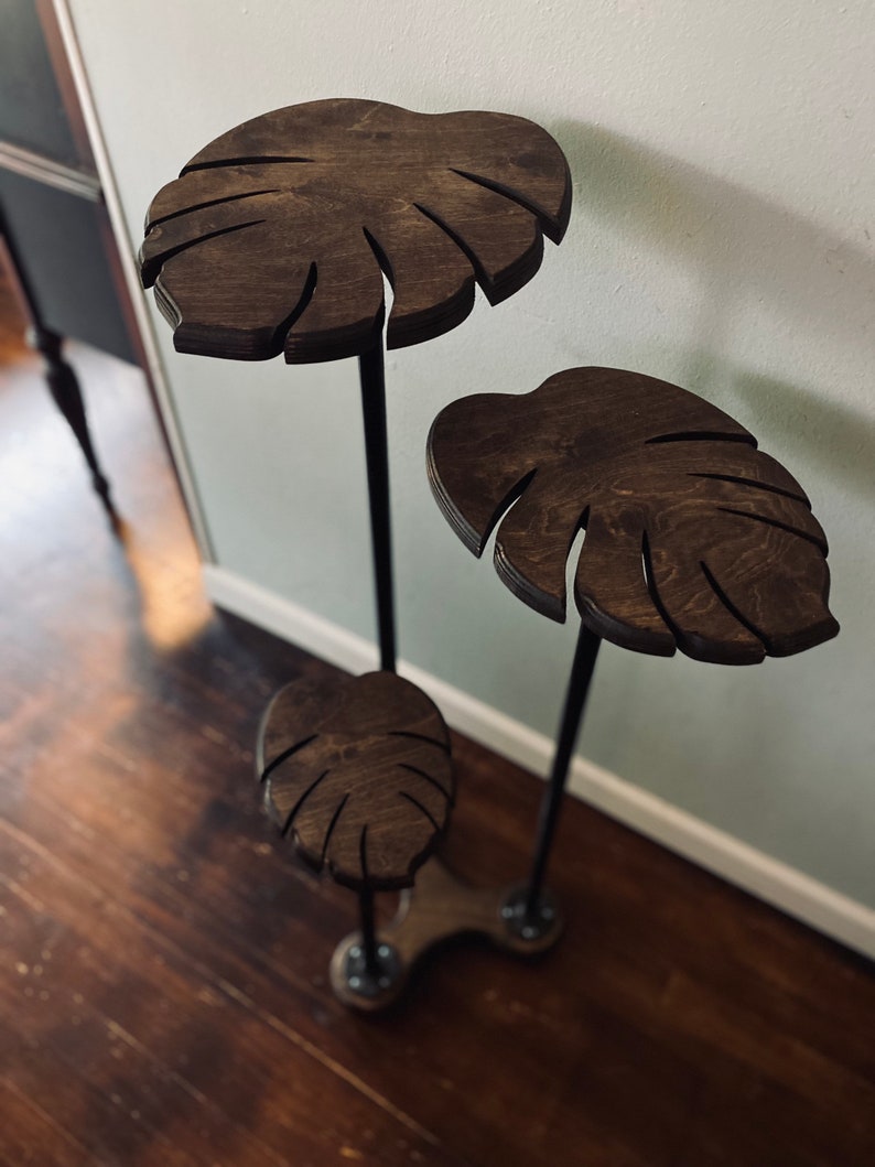 May include: A three-tier plant stand made of black metal pipes and dark brown wooden shelves shaped like monstera leaves.