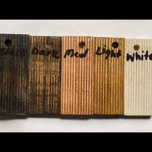 May include: A collection of five wooden samples with varying finishes. The samples are labeled "Black", "Dark", "Med", "Light", and "White". Each sample has a small hole at the top and a ridged texture.