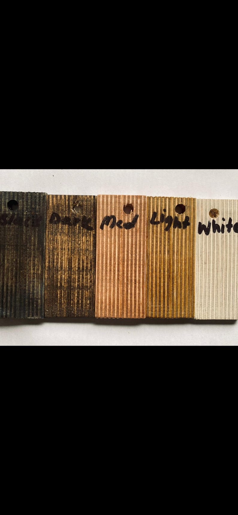 May include: A collection of five wooden samples with varying finishes. The samples are labeled "Black", "Dark", "Med", "Light", and "White". Each sample has a small hole at the top and a ridged texture.