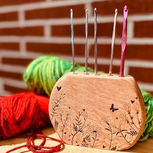 May include: Wooden crochet hook holder with a floral design and five hooks. The holder is brown and has a light wood grain. The hooks are silver and pink.