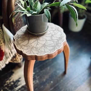 May include: A small wooden side table with three legs and a round top. The top is decorated with a floral pattern and has a plant in a gray pot sitting on it.