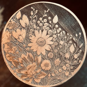 May include: A round wooden coaster featuring a laser-etched design of a floral bouquet. The flowers are in shades of brown and white, and the design is intricate and detailed.