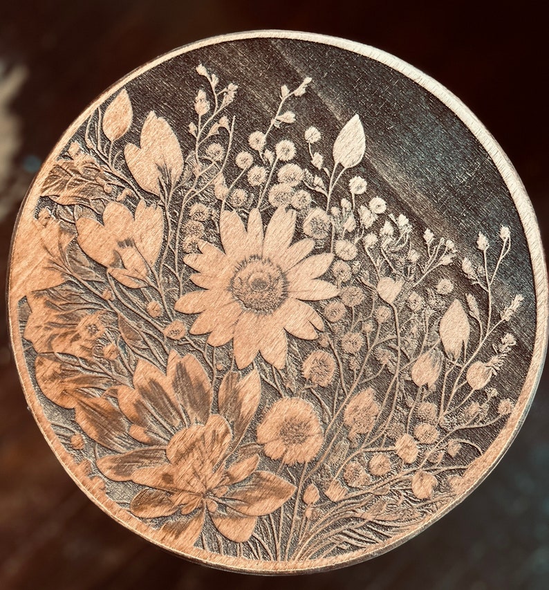 May include: A round wooden coaster featuring a laser-etched design of a floral bouquet. The flowers are in shades of brown and white, and the design is intricate and detailed.